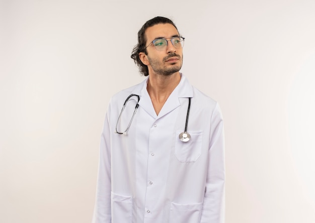 Mirando al lado joven médico con gafas ópticas vistiendo bata blanca con estetoscopio