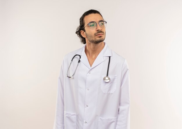 Mirando al lado joven médico con gafas ópticas vistiendo bata blanca con estetoscopio