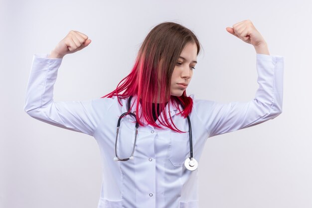 Mirando al lado joven médico vestida con estetoscopio bata médica haciendo un gesto fuerte sobre fondo blanco aislado