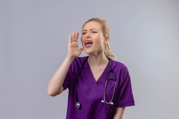 Mirando al lado joven médico vistiendo una bata médica púrpura y un estetoscopio susurra en la pared blanca aislada