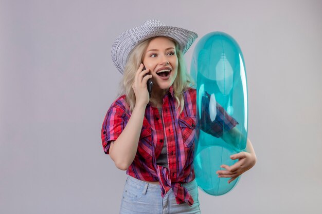 Mirando al lado joven viajera vistiendo camisa roja con sombrero sosteniendo anillo inflable habla por teléfono en la pared blanca aislada