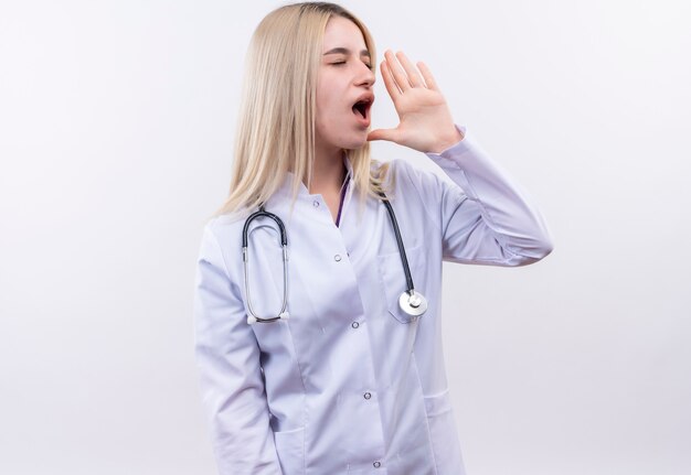 Mirando al lado médico joven rubia con estetoscopio y bata médica llamando a alguien en la pared blanca aislada