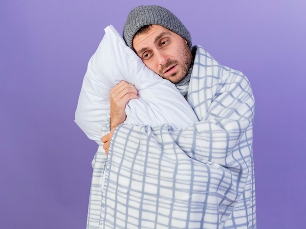 Mirando al lado molesto joven enfermo con sombrero de invierno con bufanda envuelta en una almohada abrazado a cuadros aislado sobre fondo púrpura