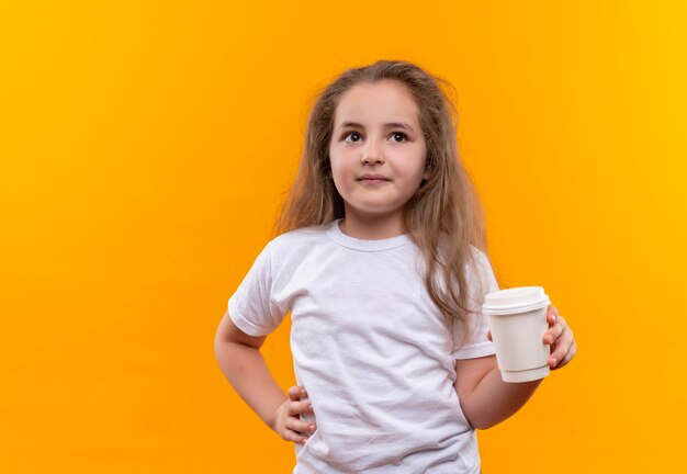 Mirando al lado de la niña de la escuela con camiseta blanca con cop de café puso su mano en la cadera en la pared naranja aislada