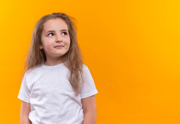 Mirando al lado de la niña de la escuela con camiseta blanca en la pared naranja aislada