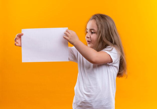 Mirando al lado de la niña de la escuela con camiseta blanca sosteniendo papel en la pared naranja aislada