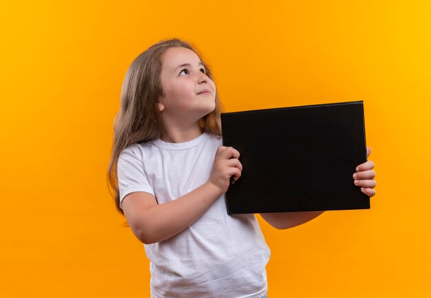 Mirando al lado de la niña de la escuela con camiseta blanca sosteniendo el portapapeles en la pared naranja aislada