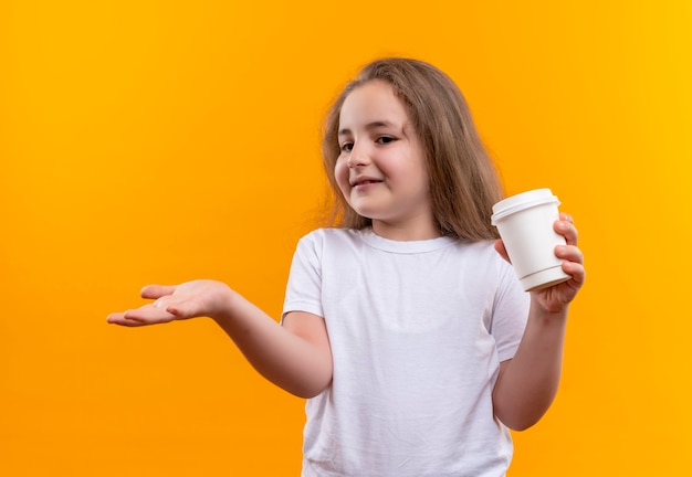 Mirando al lado de la niña de la escuela con camiseta blanca sosteniendo una taza de café en la pared naranja aislada