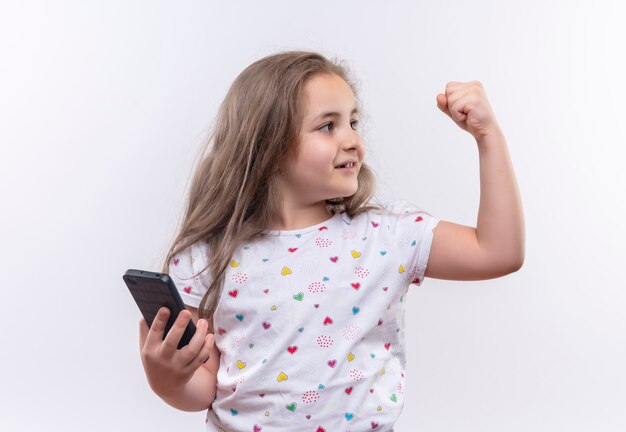 Mirando al lado de la niña de la escuela con camiseta blanca sosteniendo el teléfono mostrando un gesto fuerte en la pared blanca aislada