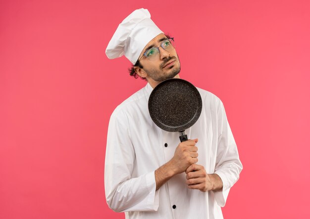 Mirando al lado pensando joven cocinero masculino vistiendo uniforme de chef y gafas sosteniendo una sartén alrededor de la barbilla