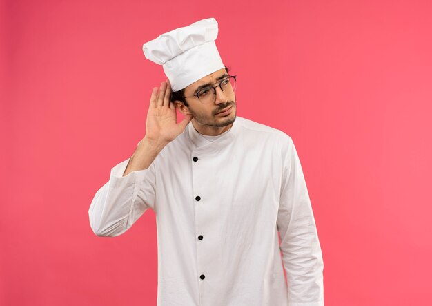 Mirando al lado pensando joven cocinero de sexo masculino vistiendo uniforme de chef y gafas mostrando gesto de escucha
