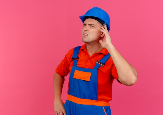 Mirando al lado pensando en el joven constructor de sexo masculino vistiendo uniforme y casco de seguridad poniendo la mano en la oreja