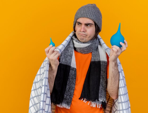 Mirando al lado pensando joven enfermo con sombrero de invierno con bufanda envuelta en cuadros sosteniendo enemas aislados sobre fondo naranja