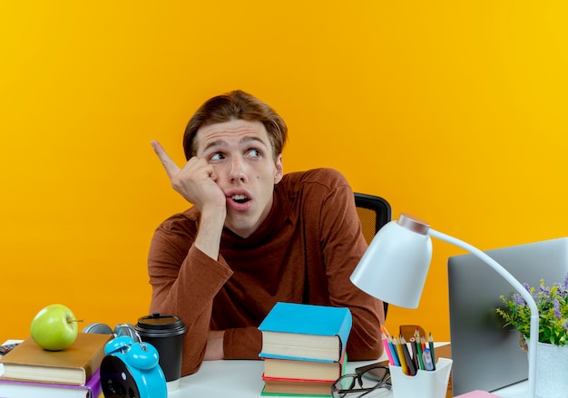 Foto gratuita mirando al lado pensando joven estudiante chico sentado en el escritorio con puntos de herramientas escolares al lado