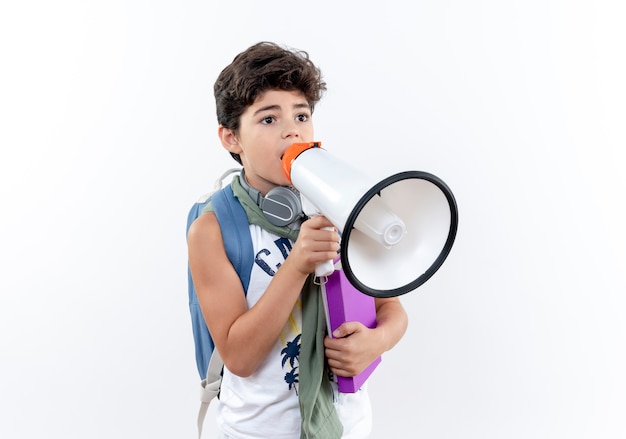 Mirando al lado pequeño colegial con mochila y auriculares sosteniendo el libro y habla por el altavoz