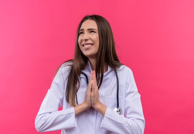 Mirando al lado preocupado joven médico vestida con estetoscopio bata médica mostrando gesto de oración en la pared rosa aislada