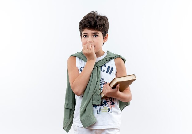 Mirando al lado preocupado pequeño colegial sosteniendo un libro y se muerde las uñas