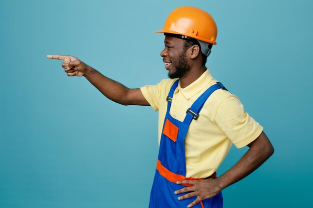 Mirando al lado sonriendo poniendo la mano en las caderas joven constructor afroamericano en uniforme aislado sobre fondo azul.