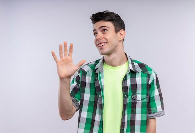 Mirando al lado sonriente joven caucásico vistiendo camisa verde mostrando cinco con la mano en la pared blanca aislada