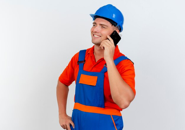 Mirando al lado sonriente joven constructor masculino vistiendo uniforme y casco de seguridad, habla por teléfono y pone la mano en la cadera