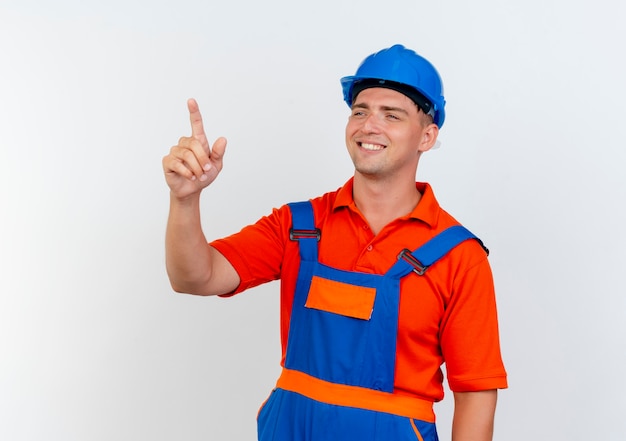 Mirando al lado sonriente joven constructor vistiendo uniforme y casco de seguridad puntos al lado