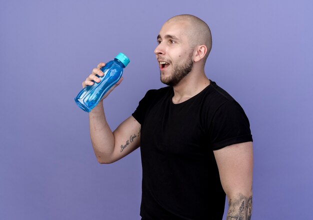 Mirando al lado sonriente joven deportivo sosteniendo una botella de agua aislada en la pared púrpura