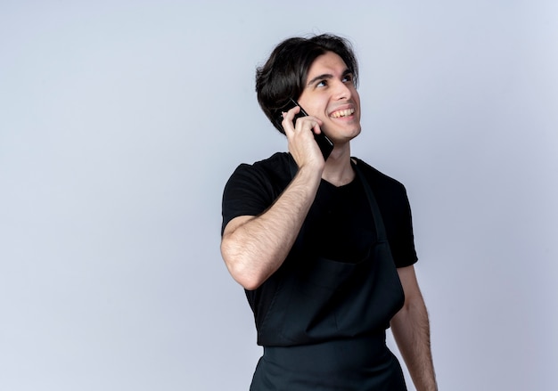 Mirando al lado sonriente joven guapo barbero en uniforme habla por teléfono