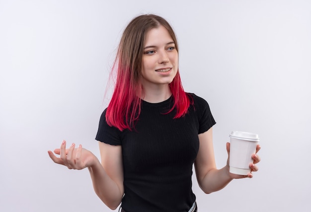 Mirando al lado sonriente joven hermosa mujer vistiendo camiseta negra sosteniendo una taza de café en la pared blanca aislada