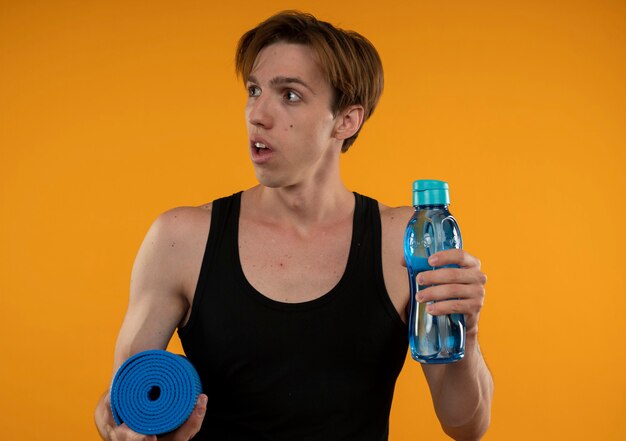 Mirando al lado sorprendido joven deportivo sosteniendo una estera de yoga con una botella de agua aislada en la pared naranja