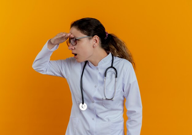 Mirando al lado sorprendido joven doctora vistiendo bata médica y estetoscopio con gafas mirando a distancia con la mano aislada en la pared naranja