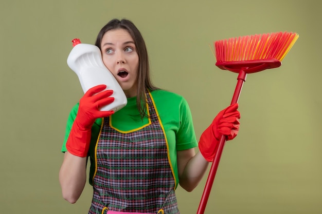 Foto gratuita mirando al lado sorprendido limpieza joven vistiendo uniforme en guantes rojos sosteniendo trapeador poner agente de limpieza en la mejilla en su mano en la pared verde aislada