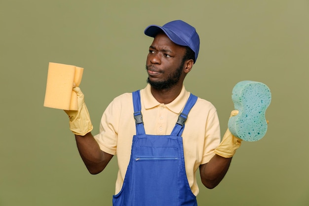 Mirando al lado sosteniendo esponjas de limpieza joven limpiador afroamericano en uniforme con guantes aislados en fondo verde