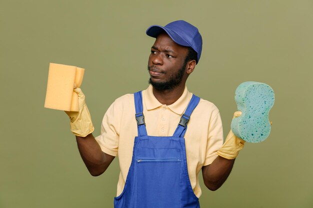 Mirando al lado sosteniendo esponjas de limpieza joven limpiador afroamericano en uniforme con guantes aislados en fondo verde
