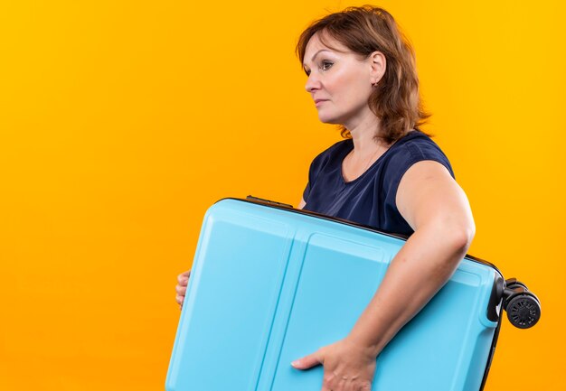 Mirando al lado viajero de mediana edad mujer sosteniendo maleta sobre fondo amarillo aislado