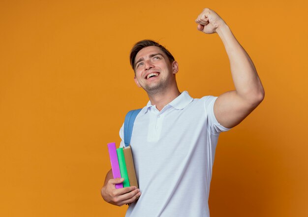 Mirando hacia arriba alegre joven apuesto estudiante vistiendo bolsa trasera levantando el puño aislado en la pared naranja