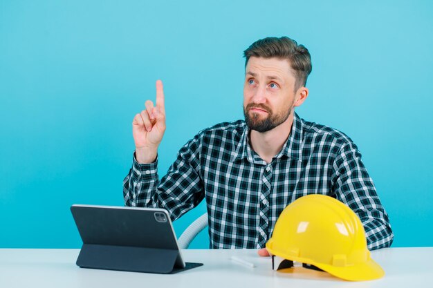 Mirando hacia arriba, el arquitecto está apuntando hacia arriba con el dedo índice sentado frente a la tableta sobre fondo azul.