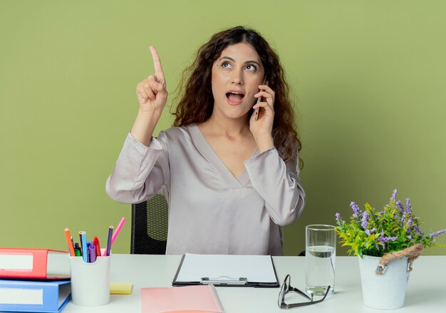 Mirando hacia arriba complacida joven oficinista bastante femenina sentada en el escritorio con herramientas de oficina habla por teléfono y apunta hacia arriba aislado sobre fondo verde oliva con espacio de copia