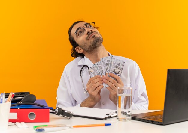 Foto gratuita mirando hacia arriba complacido joven médico con gafas médicas vistiendo bata médica con estetoscopio sentados en el escritorio