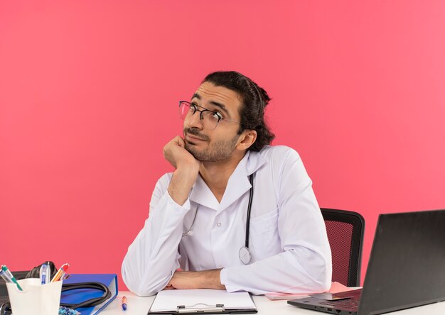 Mirando hacia arriba complacido joven médico con gafas médicas vistiendo bata médica con estetoscopio sentados en el escritorio