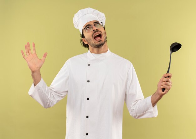 Mirando hacia arriba enojado joven cocinero vistiendo uniforme de chef y gafas sosteniendo la cuchara y extendiendo la mano