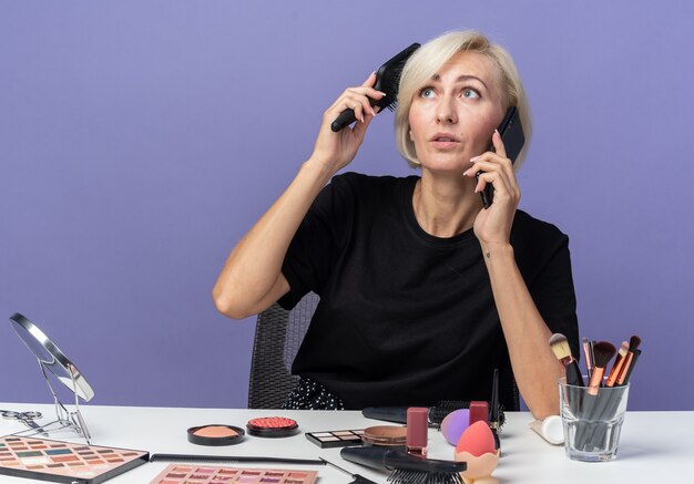 Mirando hacia arriba la hermosa joven se sienta a la mesa con herramientas de maquillaje habla por teléfono peinando el cabello aislado en la pared azul