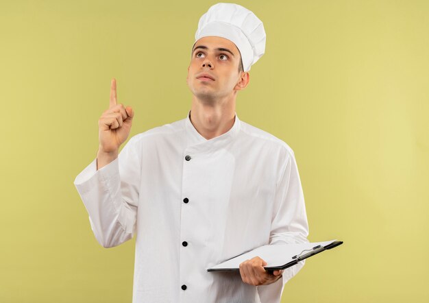 Mirando hacia arriba el joven cocinero con uniforme de chef apunta con el dedo hacia arriba y sosteniendo el portapapeles en la pared verde aislada