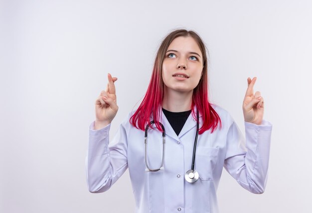 Mirando hacia arriba la joven doctora vestida con estetoscopio bata médica cruzando los dedos sobre fondo blanco aislado