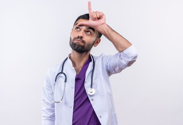 Mirando hacia arriba el joven médico con estetoscopio bata médica apunta hacia arriba sobre fondo blanco aislado