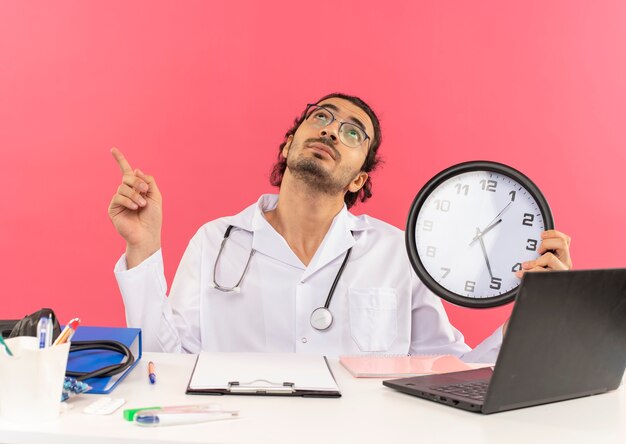 Mirando hacia arriba el joven médico con gafas médicas vistiendo bata médica con estetoscopio sentados frente al escritorio
