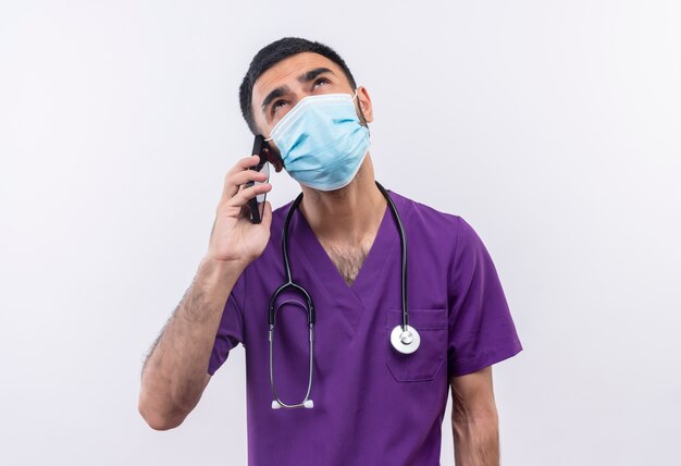 Mirando hacia arriba el médico varón joven vistiendo ropa de cirujano púrpura y estetoscopio máscara médica habla por teléfono sobre fondo blanco aislado