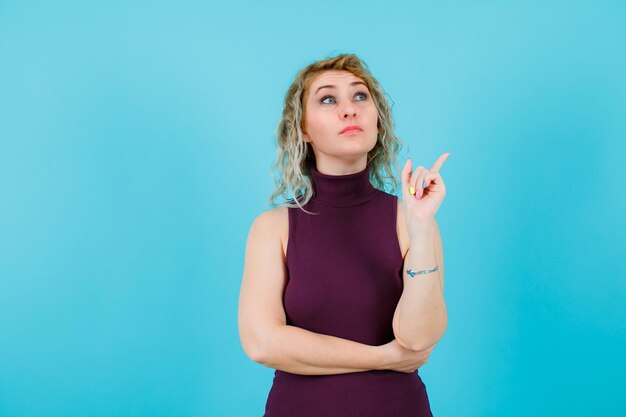 Mirando hacia arriba, la mujer rubia está apuntando hacia arriba con el dedo índice sobre fondo azul.