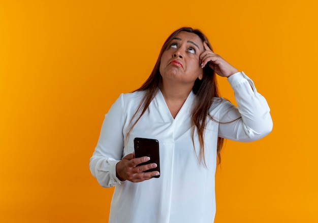 Mirando hacia arriba pensando casual mujer caucásica de mediana edad sosteniendo el teléfono y poniendo el dedo en la frente