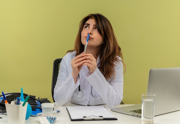 Mirando hacia arriba pensando en doctora de mediana edad vistiendo bata médica con estetoscopio sentado en el escritorio trabajar en una computadora portátil con herramientas médicas poniendo la pluma en la boca en la pared verde con espacio de copia