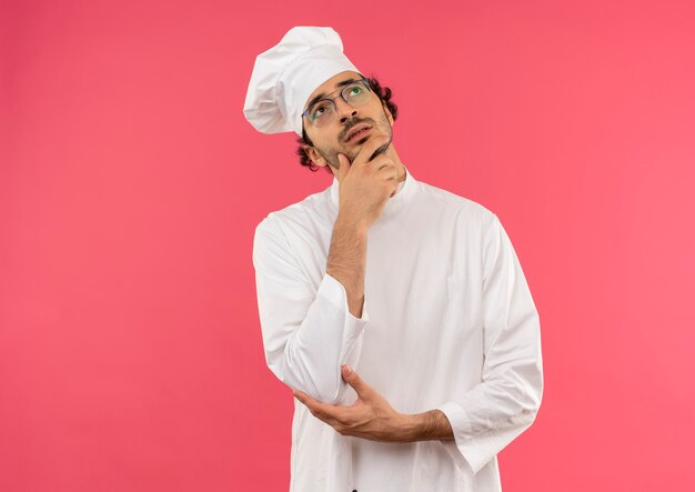 Mirando hacia arriba pensando joven cocinero vistiendo uniforme de chef y gafas poniendo la mano en la barbilla